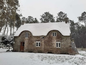 A stone house with a snow-covered roof and a wooden door, surrounded by trees and a snowy landscape.