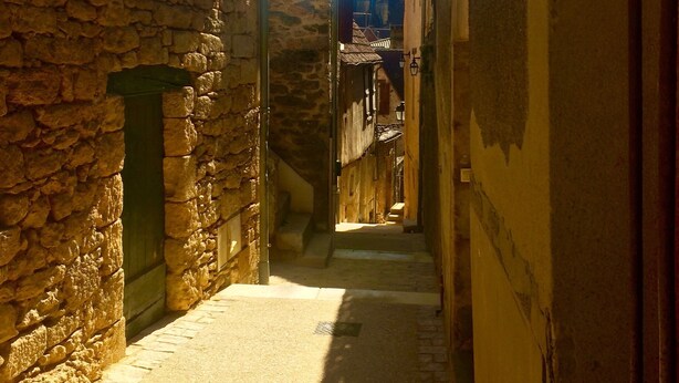 Une ruelle étroite aux murs de pierre et aux portes en bois, qui mène à un immeuble dont on aperçoit un escalier.