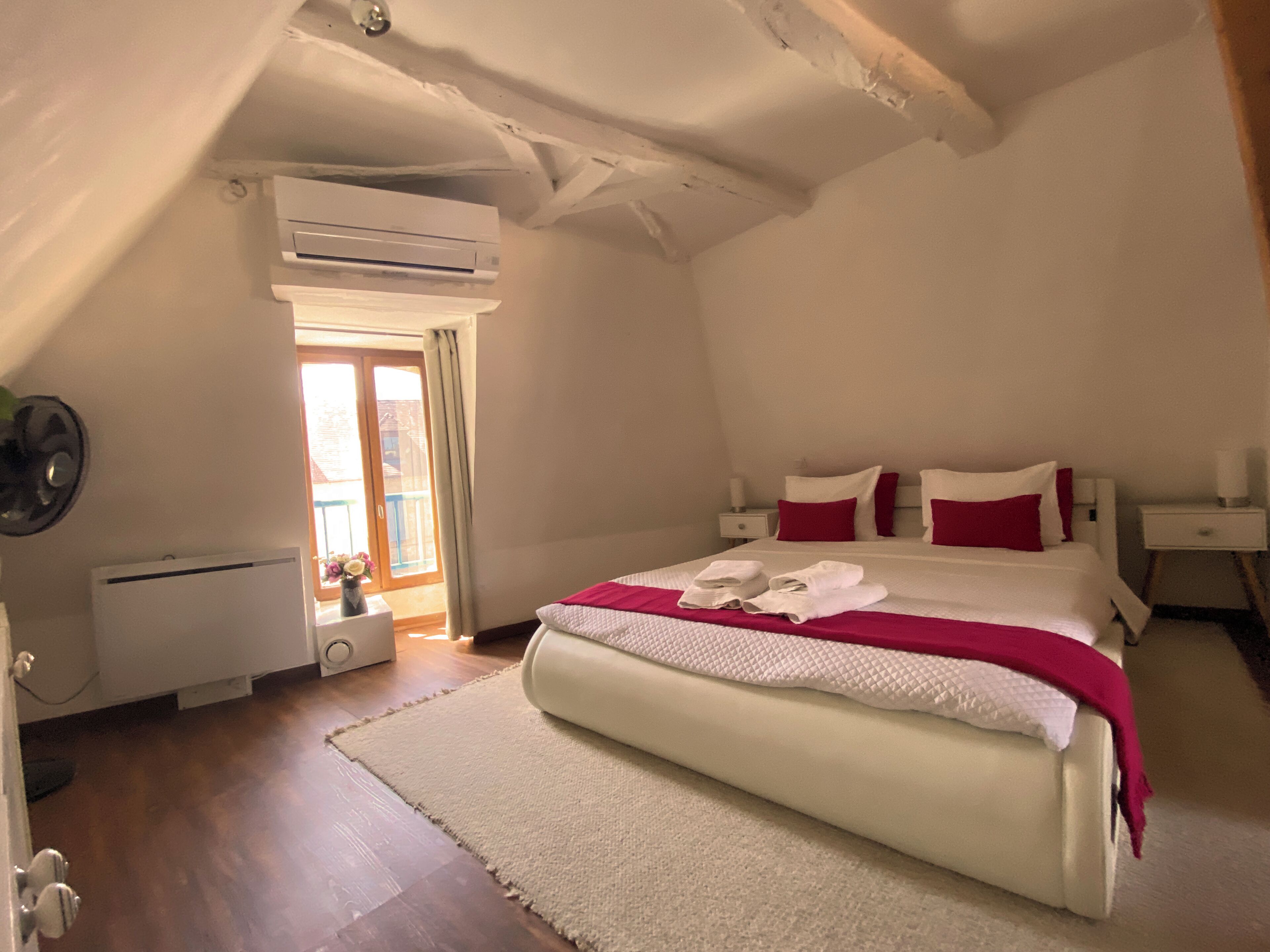 A bedroom with a bed, a bedside table, a nightstand, an air conditioning unit, a window with a view, and a door.