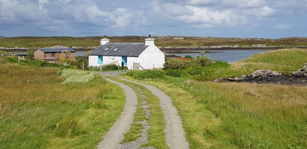 Un chemin de campagne mène à une maison blanche aux bordures bleues et au toit gris, entourée de champs verdoyants et bordée d’un plan d’eau.