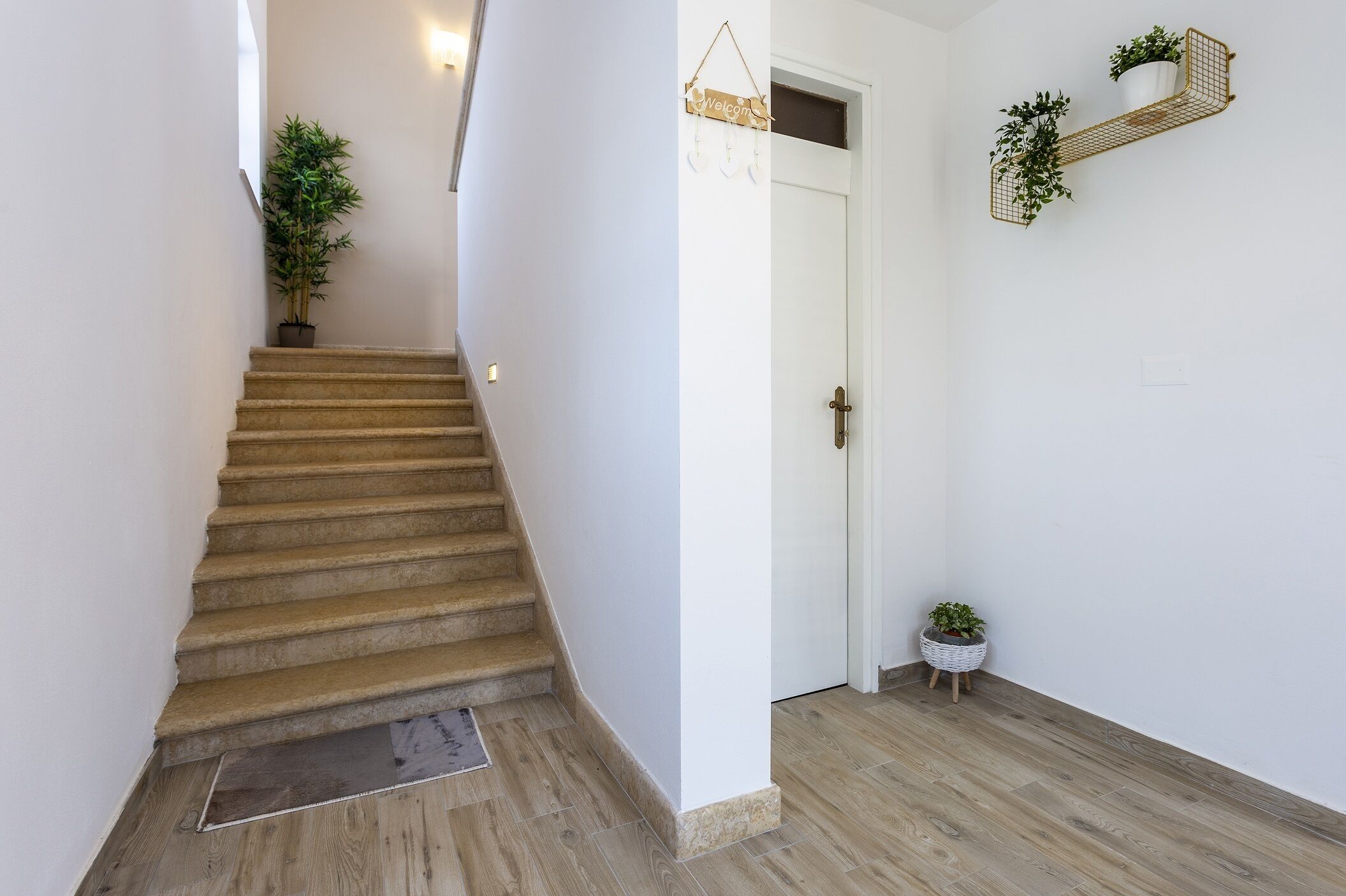 En trappe fører op til en dør med et velkomstskilt, en potteplante på repos og en lille plante på gulvet.