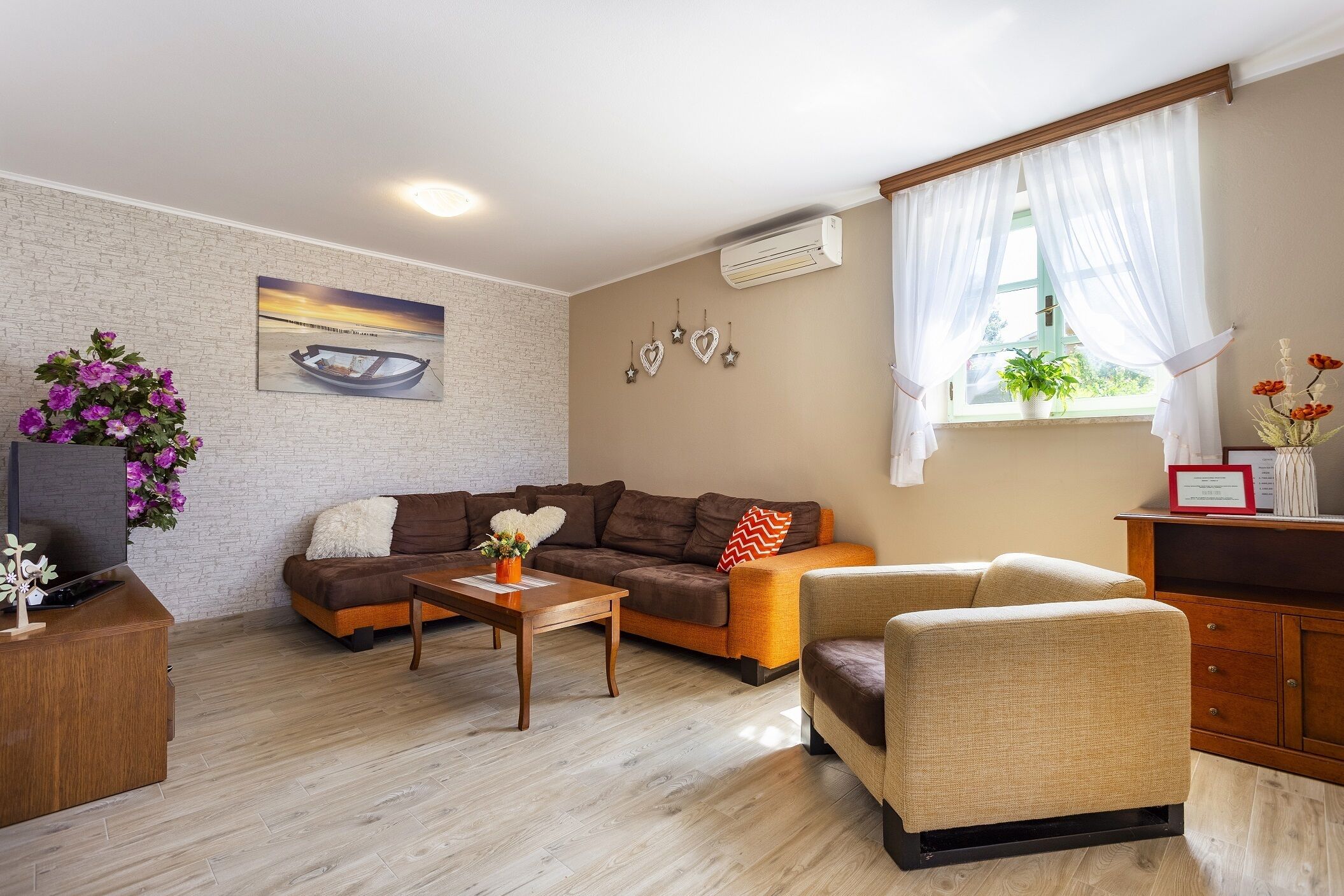A cozy living room with a brown sofa, a beige armchair, and a wooden coffee table. There is a TV on a wooden stand, a potted plant, and a painting on the wall.