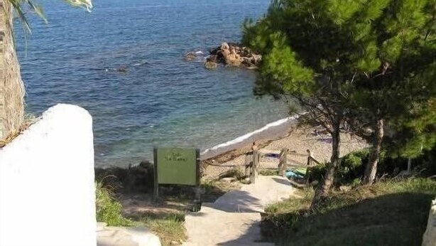 A coastal view with a clear path leading to a beach, a sign indicating a location, and a rocky outcrop in the water. - Sun, sea, calm and space in a finca 6.5 ha, Casita Tres (El Perelló)