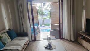Un salon comprenant un canapé, une table basse et un meuble pour la télévision. Une porte-fenêtre coulissante donne accès à un espace extérieur avec une piscine et du mobilier.