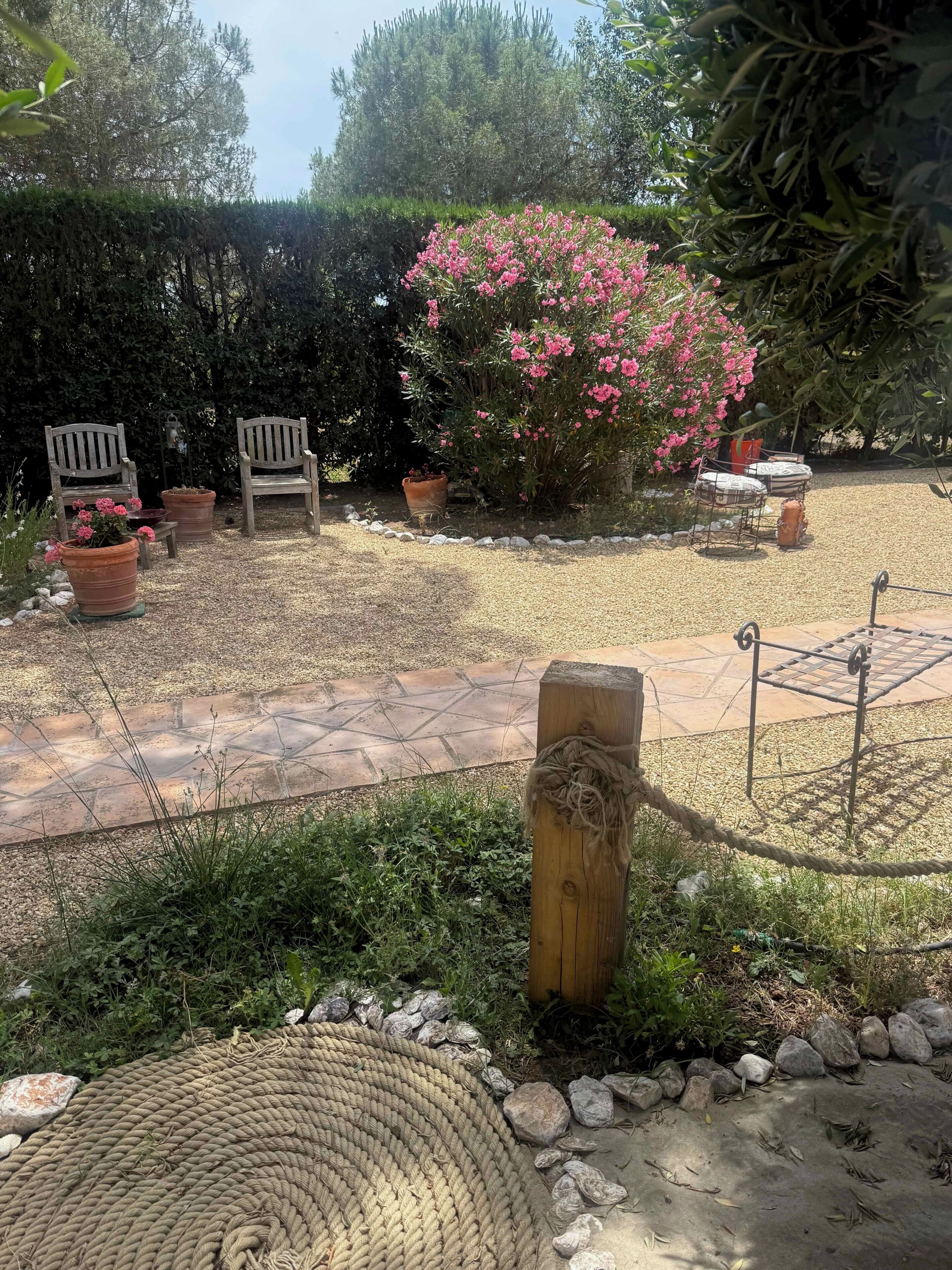 Een tuinpatio met een pad van stenen, houten stoelen, plantenpotten en een touwbordes.