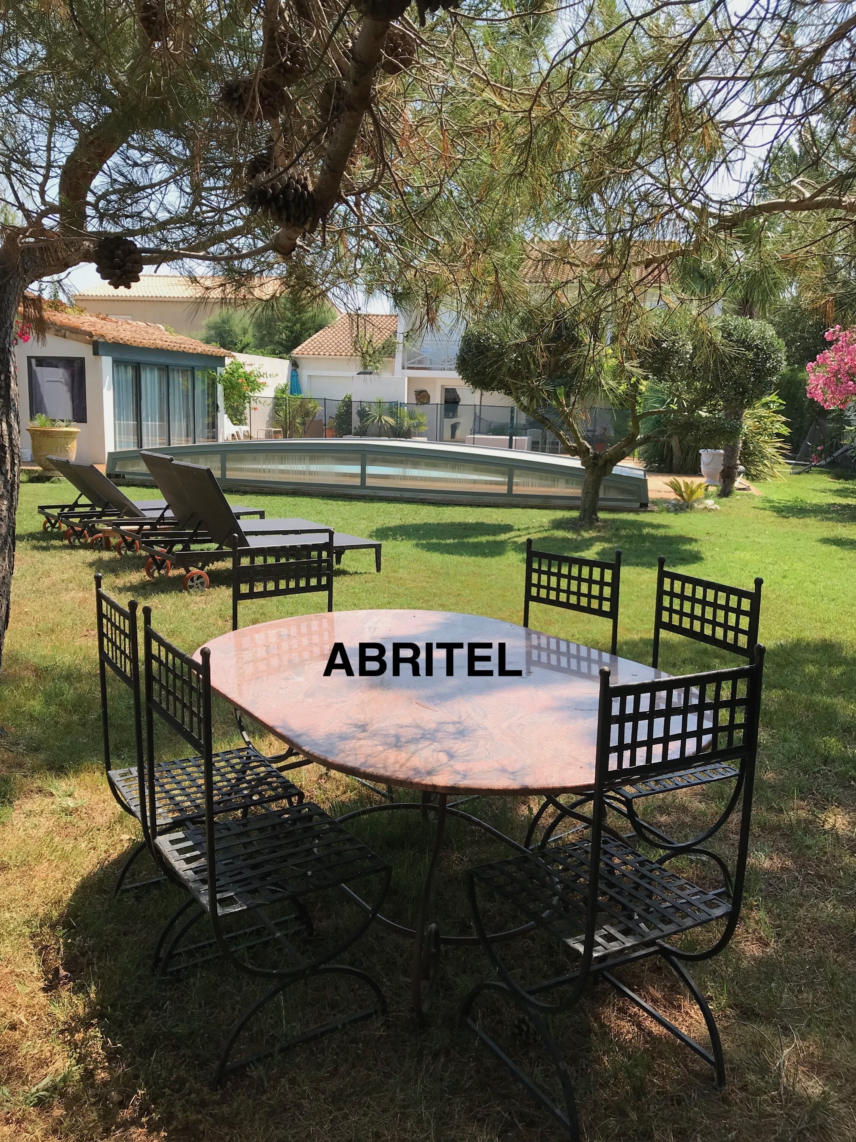 A round metal table with four chairs in a grassy outdoor area, with a pool and buildings in the background.