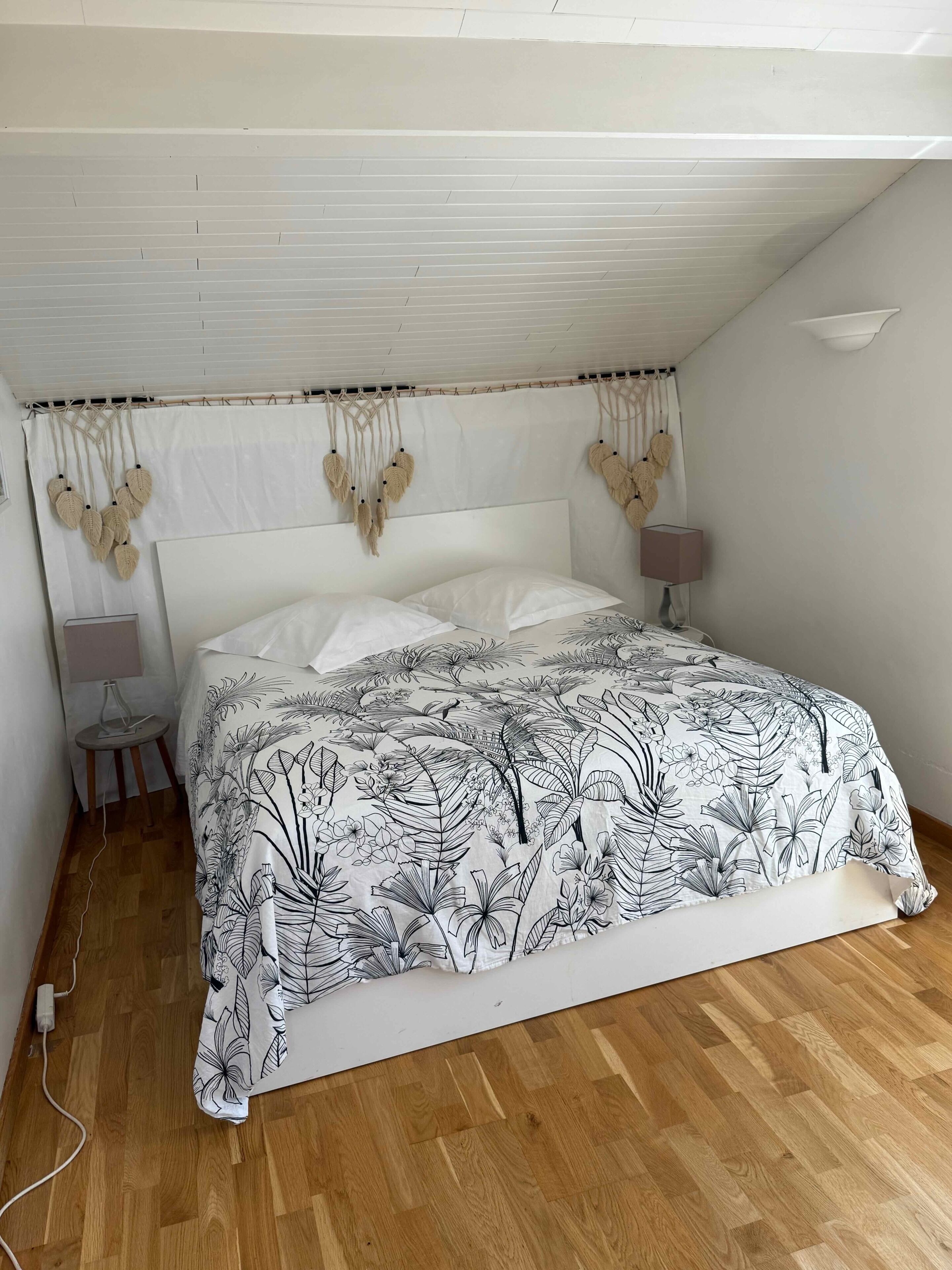Un dormitorio con una cama, una mesita de noche, una lámpara, un enchufe y un techo con panel blanco.