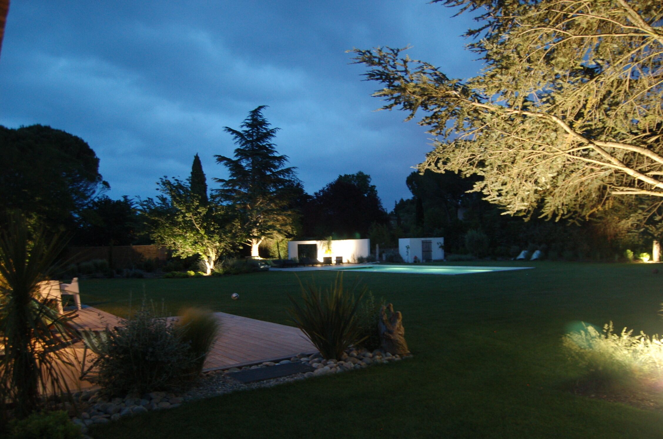 A well-lit backyard with a swimming pool, surrounded by trees and a patio area.