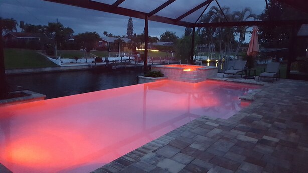 An outdoor swimming pool with a red-lit edge, surrounded by a covered patio area with chairs and a view of a residential neighborhood. - Villa Alani -- Luxurious south-facing dream villa directly on the canal / river connection (Cape Coral)