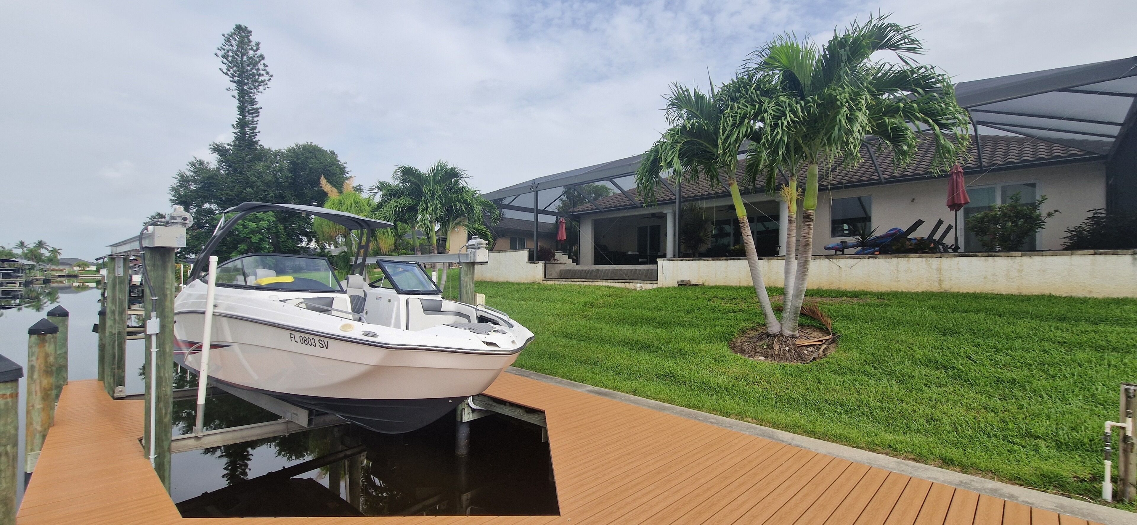A boat docked at a wooden pier with a house and palm trees in the background.