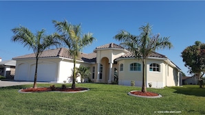 A two-story house with a beige exterior, a brown tiled roof, and a well-maintained lawn with palm trees. - Villa Alani -- Luxurious south-facing dream villa directly on the canal / river connection (Cape Coral)
