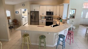 Une cuisine moderne avec des meubles blancs, un îlot central doté de tabourets, des appareils en acier inoxydable et un coin repas avec une table et des chaises.