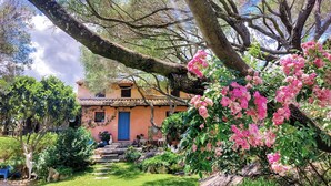 A rustic house with a tiled roof, surrounded by lush greenery and pink flowers. - Garden Villa near Costa Smeralda (Olbia)