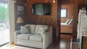 A cozy cabin interior with a wooden ceiling, a flat-screen TV mounted on the wall, a beige sofa with patterned cushions, a small kitchen area with a refrigerator, and a wooden deck visible through a sliding door. - Cozy Waterfront Cottage -- No Wake Cove, Great Views, New Private Floating Dock (Huddleston)