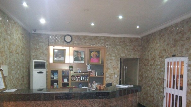 A reception area with a marble counter, framed photographs, a clock, and a door with vertical bars. - Hotel Codial Exclusive (Enugu)
