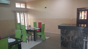 A dining area with a round table and green chairs, a small bar area with a marble countertop, and a window with blinds. - Hotel Codial Exclusive (Enugu)