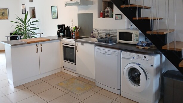 Une cuisine moderne avec des armoires blanches, un four encastré, un four à micro-ondes et un lave-linge. On y trouve un comptoir en bois et un escalier orné d’une pancarte « BIENVENUE ».