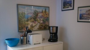 Une petite cuisine équipée d’un grille-pain, d’une machine à café et d’un bol bleu posé sur un meuble blanc. Deux tableaux encadrés ornent le mur.