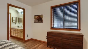 Een slaapkamer met een houten vloer, een bed met een beddegoed met bladmotief, een dressoir, een raam met jaloezieën, een spiegel en een zichtbare badkamer door een open deur.