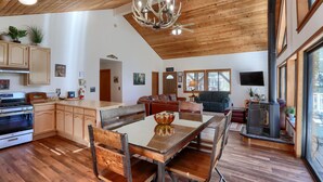 Een ruime woonkamer met een houten plafond, een eettafel met stoelen, een open haard en een aan de muur bevestigde televisie.