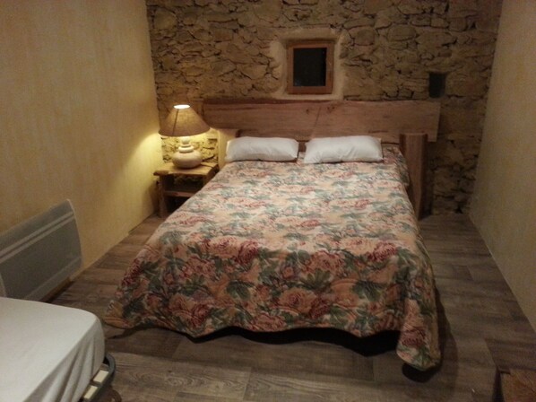 Una habitación pequeña con cama, mesita de noche, lámpara y pared de piedra.