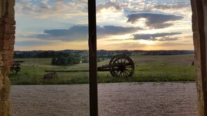 A view through a window with a rustic brick wall, a wooden table, and a large wooden wheel in a grassy field during sunset.