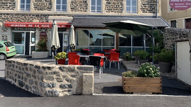 Ein Steinhaus mit einem Restaurant namens "Auberge de la Tour", das Sitzgelegenheiten im Freien mit roten Stühlen und Sonnenschirmen bietet.