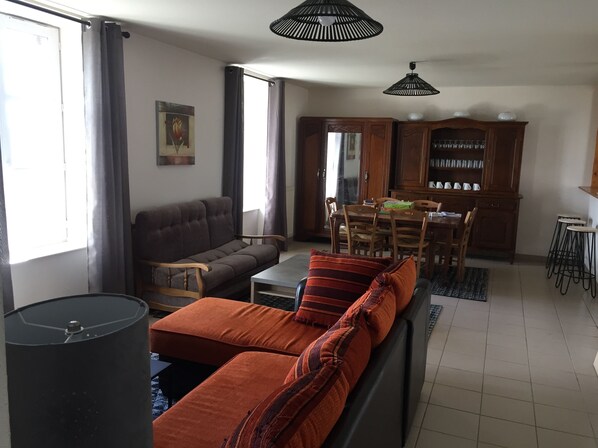 A living room with a sofa, a dining table, and chairs. There is a cabinet with glassware and a small table with stools.