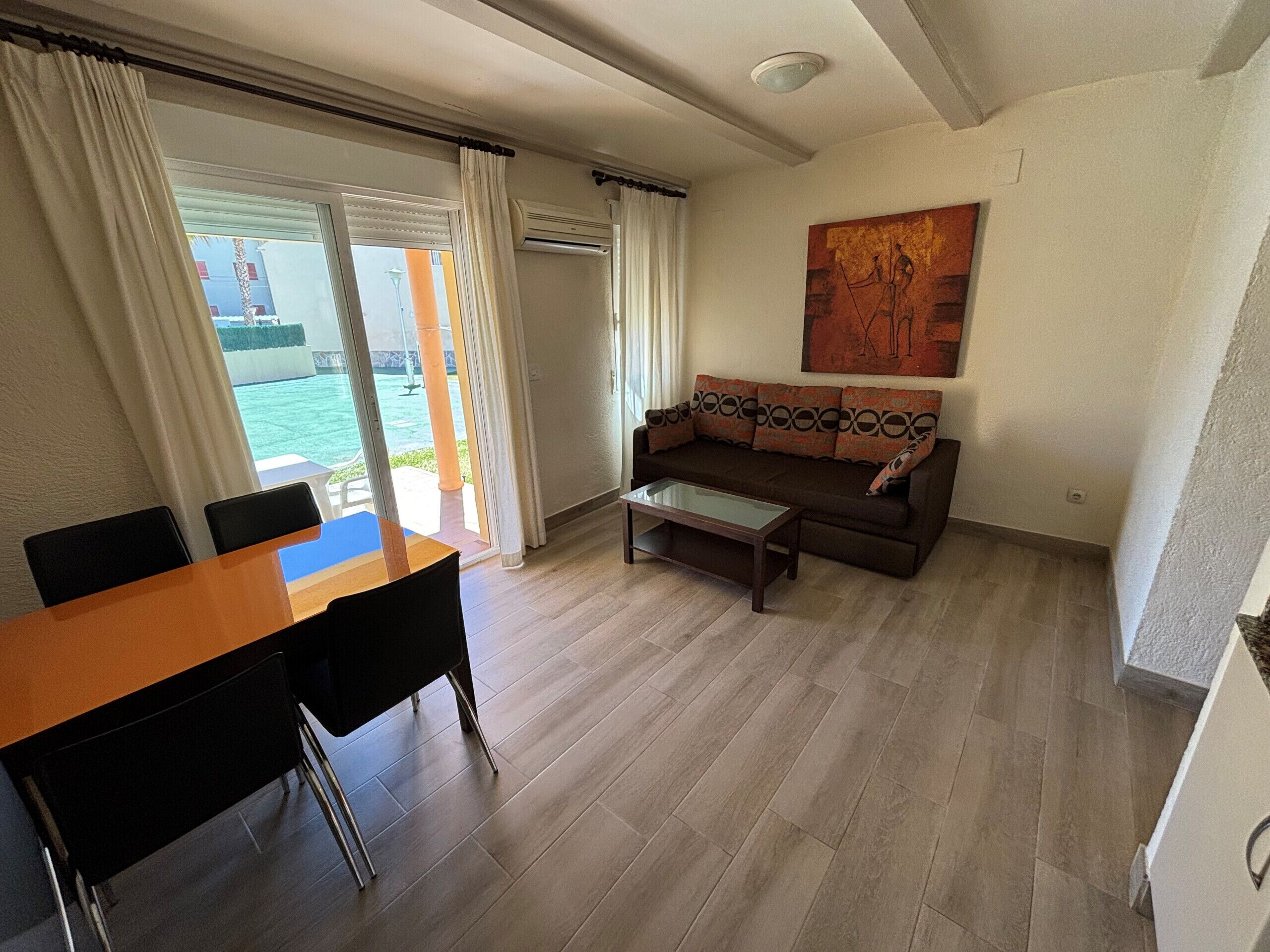 Un salon comprenant un canapé, une table basse et une table à manger avec des chaises. On peut admirer la piscine depuis l’intérieur.
