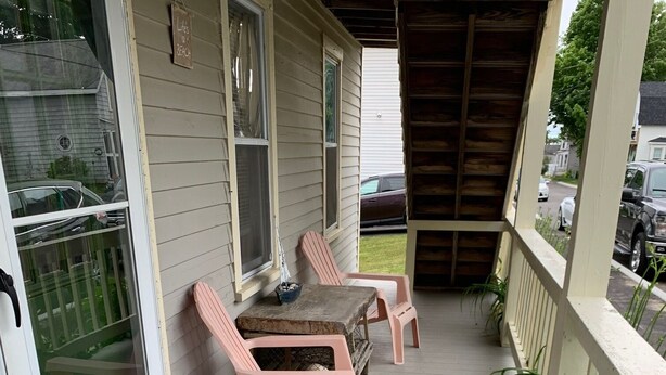 A covered porch with two pink chairs and a table, a wooden deck, and a white door with a window. - 3 Bdr Vacation Rental - Walk To The Beach (Old Orchard Beach)