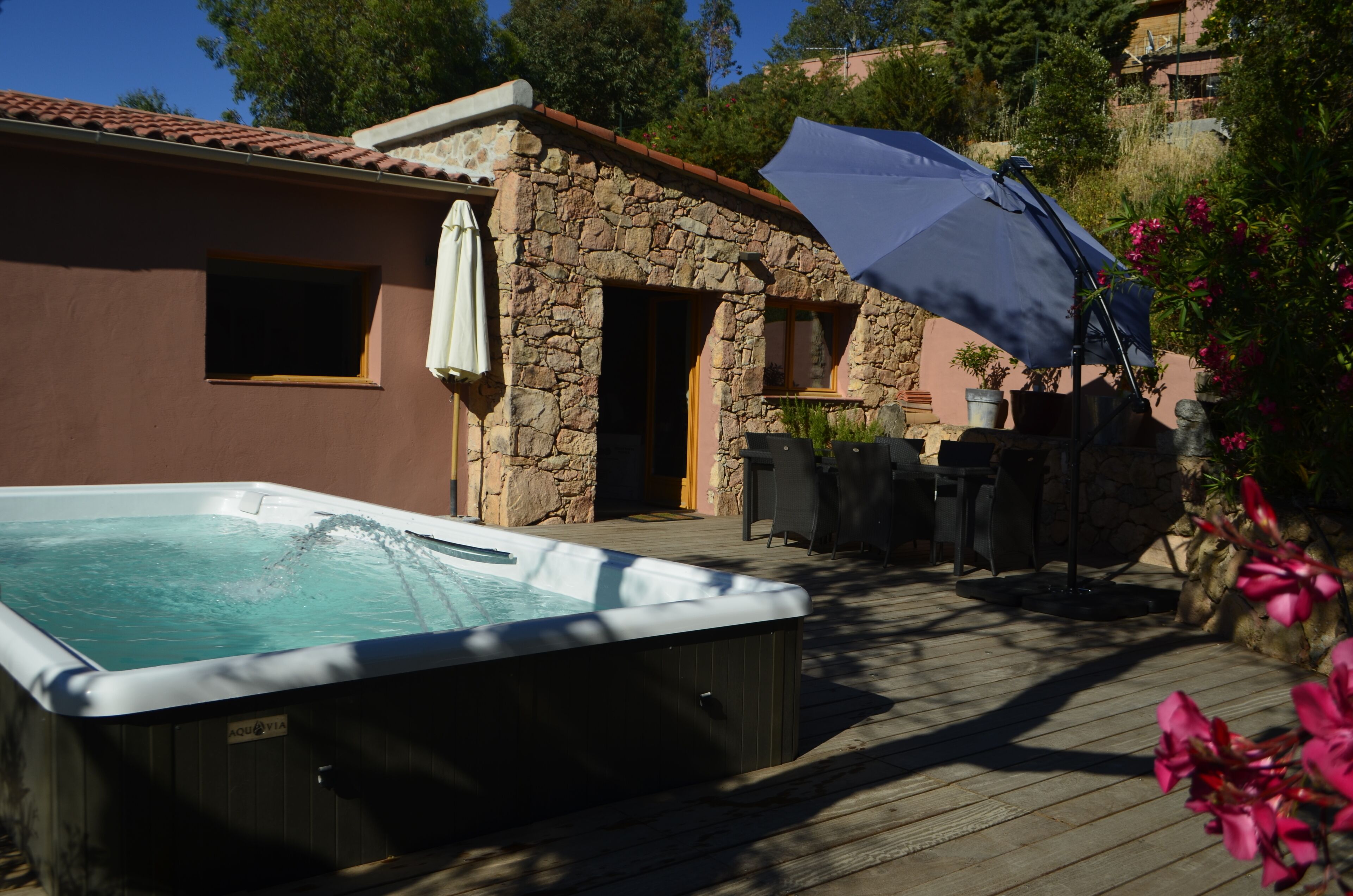 A hot tub in front of a stone building with a wooden deck, a patio area, and a white umbrella.