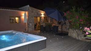 A hot tub on a wooden deck, a stone building with a door, and outdoor furniture under an umbrella.