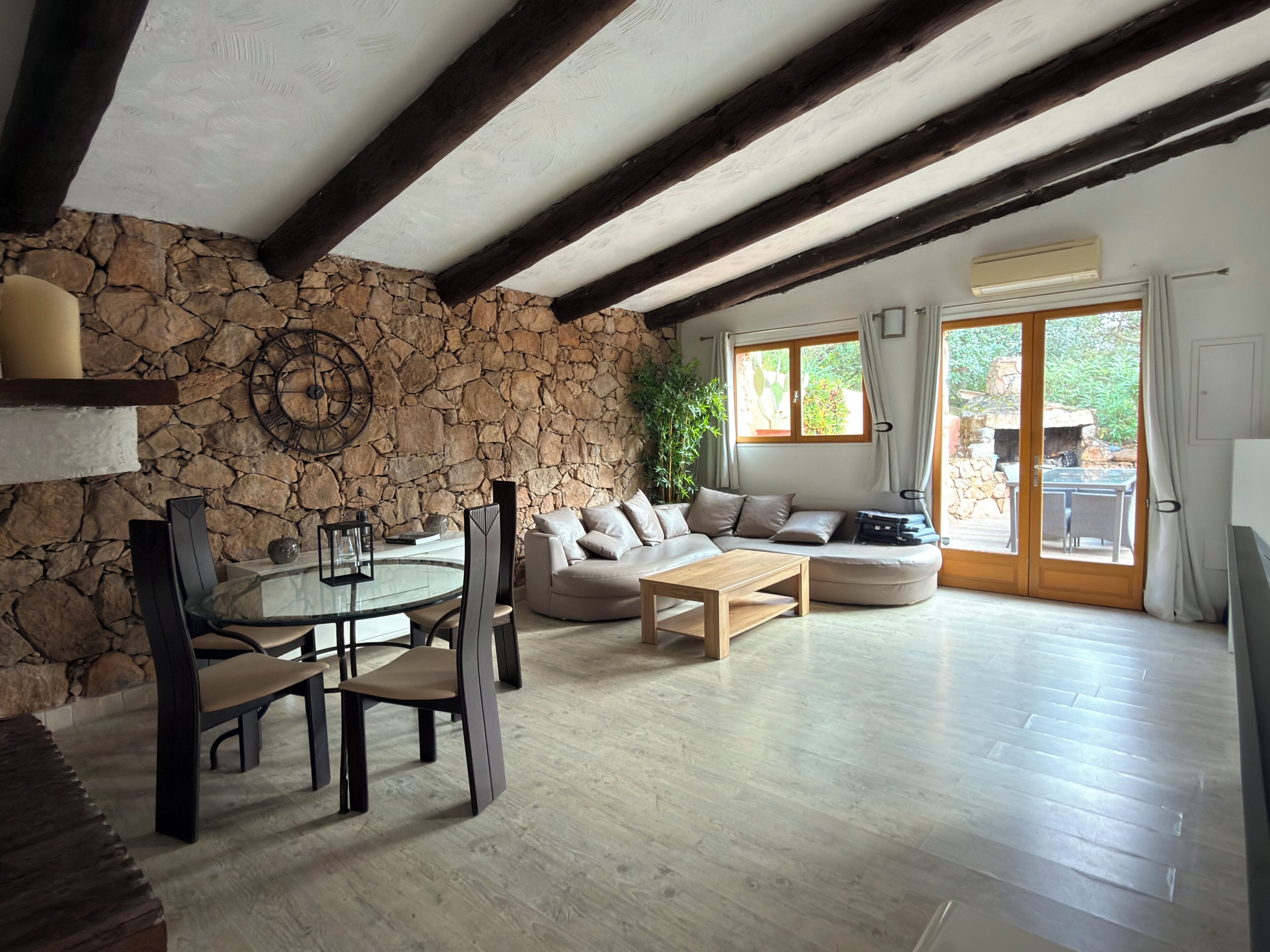 Un salon avec un mur en pierre, des poutres apparentes, une table ronde entourée de chaises, un canapé et une vue sur l’extérieur, visible à travers une porte vitrée.