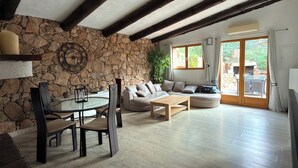 A living room with a stone wall, wooden beams, a round table with chairs, a sofa, and a view through a glass door.