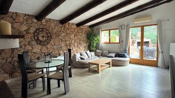 Un salon avec un mur en pierre, des poutres apparentes, une table ronde entourée de chaises, un canapé et une vue sur l’extérieur, visible à travers une porte vitrée.