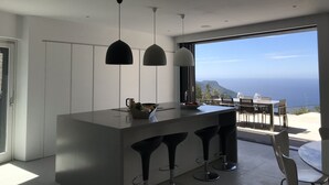 A modern kitchen with a central island, bar stools, pendant lights, and a view of the ocean. - STUNNING LUXURY HOME WITH SEA, MOUNTAIN & SUNSET VIEWS. Private pool & terraces (Valldemossa, Mallorca)