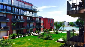 Un immeuble à plusieurs étages doté de balcons, une cour avec de la pelouse, des plantes en pot et une vue sur une étendue d’eau.