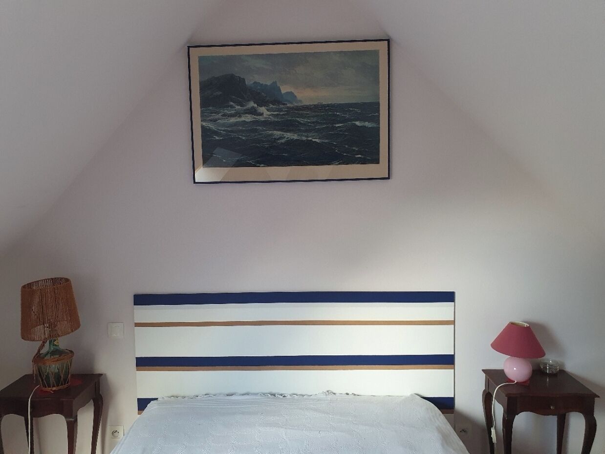 Une chambre à coucher avec un lit, une tête de lit à rayures horizontales, un tableau représentant un paysage marin et deux tables de chevet avec des lampes.