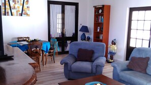A living room with a wooden coffee table, two blue armchairs, a wooden side table, a bookshelf, and a dining area with a table and chairs. - the cottages of castera NORMANDIE (Aire-Sur-L'adour)