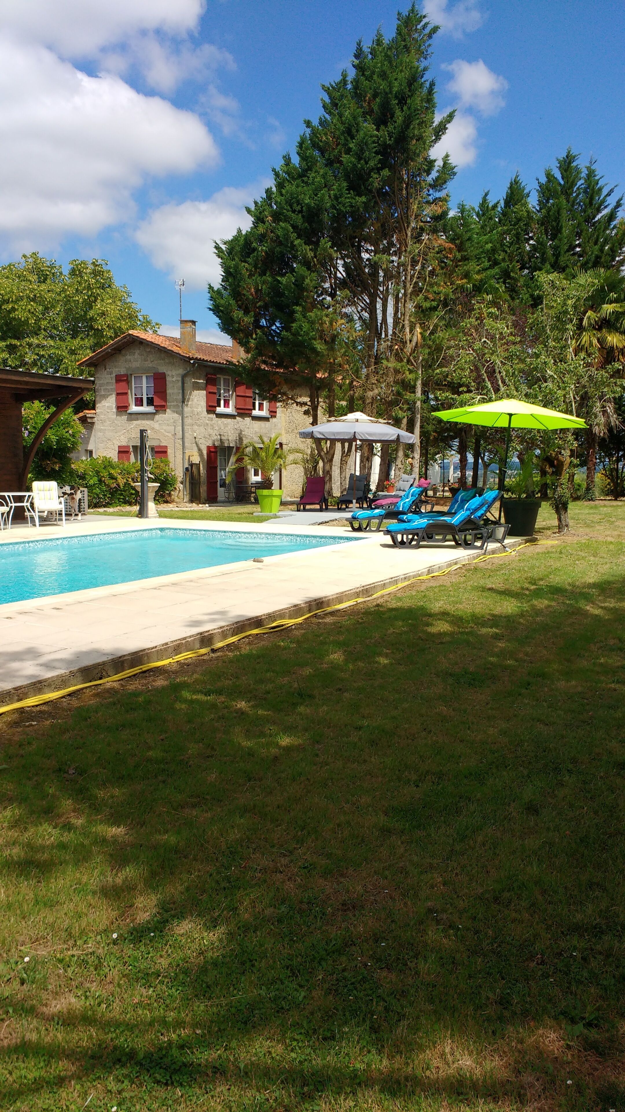 Une piscine entourée d’arbres, avec des chaises longues et des parasols, et un bâtiment en pierre aux volets rouges.