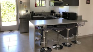 Une cuisine moderne équipée d’un réfrigérateur en acier inoxydable, d’un four encastré, d’un four à micro-ondes et d’une machine à café. On y trouve quatre tabourets de bar disposés autour d’un îlot central.
