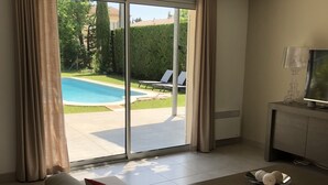 Un salon moderne avec un canapé, une table basse et un téléviseur posé sur un meuble. De grandes portes-fenêtres coulissantes donnent accès à une piscine et à un espace de détente extérieur.