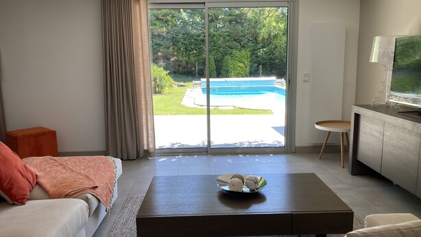 Un salon moderne avec un canapé, une table basse et un téléviseur posé sur un meuble. Une porte-fenêtre coulissante donne accès à un espace piscine.