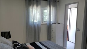 Ein Schlafzimmer mit Bett, Nachttischen, einem Fenster mit Vorhängen, einer Tür und Blick ins Freie.