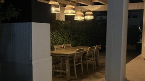 Ein Essbereich im Freien mit einem Holztisch und Stühlen, umgeben von einer niedrigen Mauer und einem überdachten Sitzbereich mit hängenden Lampen.