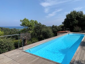 Une piscine rectangulaire avec une terrasse en bois, entourée d’arbres et offrant une vue sur l’océan.