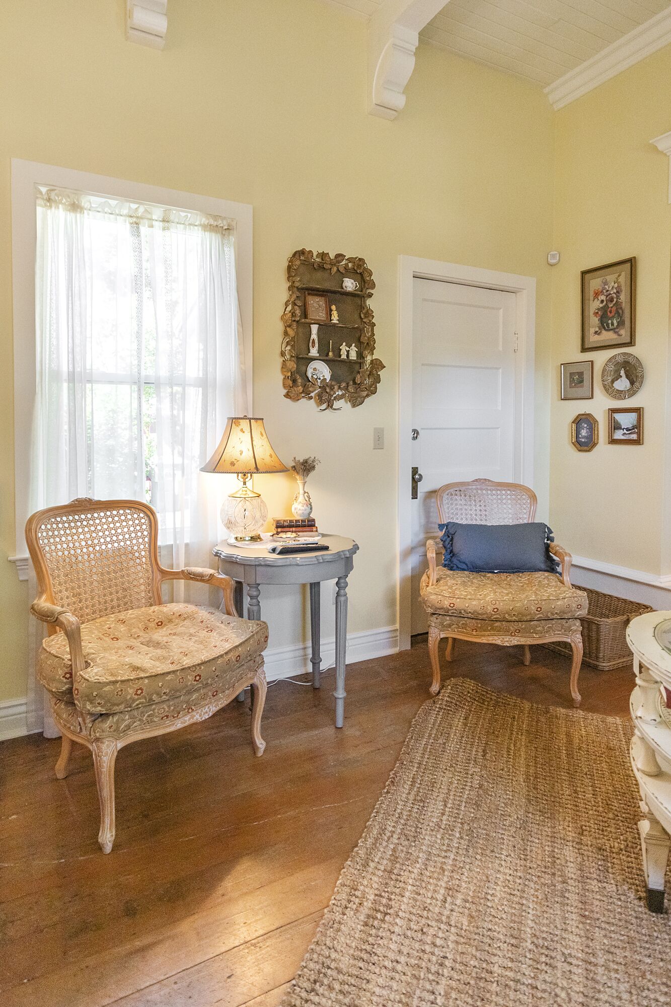 Una habitación con dos sillones antiguos de mimbre, una mesita redonda con una lámpara y una puerta con un espejo decorativo encima.