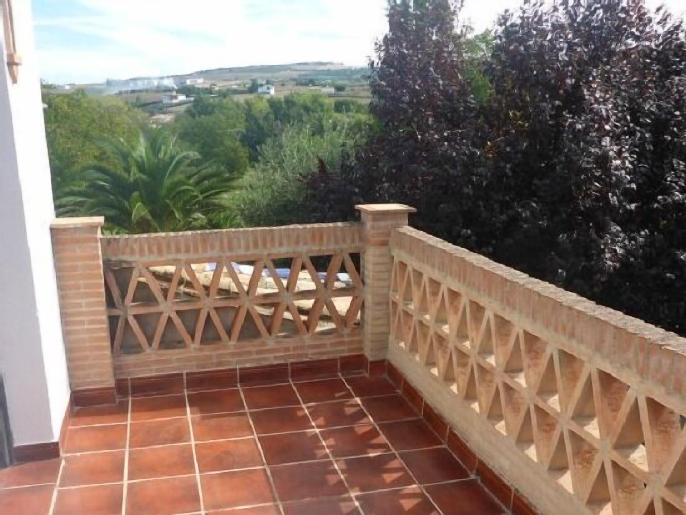 Un balcón con baldosas de terracota, barandilla de madera retorcida y vistas a zonas verdes y edificios a lo lejos.