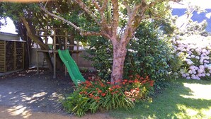 Un albero con uno scivolo, circondato da un giardino con fiori rossi e un siepe fiorita di rosa.
