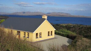 Ein gelbes Haus mit grauem Dach, einem Schotterweg und Blick auf das Meer und die Berge im Hintergrund.